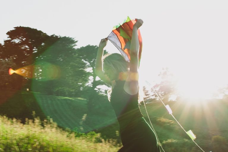 Musician Anya Anastasia dancing in a sunny outdoor field holding a colorful fabric overhead, backlit by golden sunlight with lens flare.
